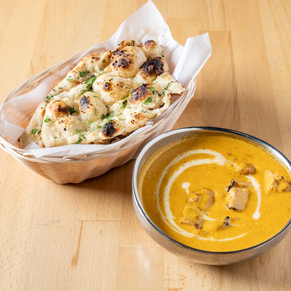 A basket of naan bread with cilantro next to a bowl of butter chicken on a wooden table.