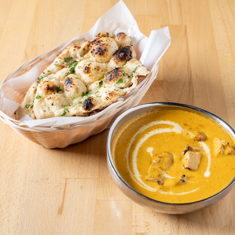 A basket of naan bread with cilantro next to a bowl of butter chicken on a wooden table.