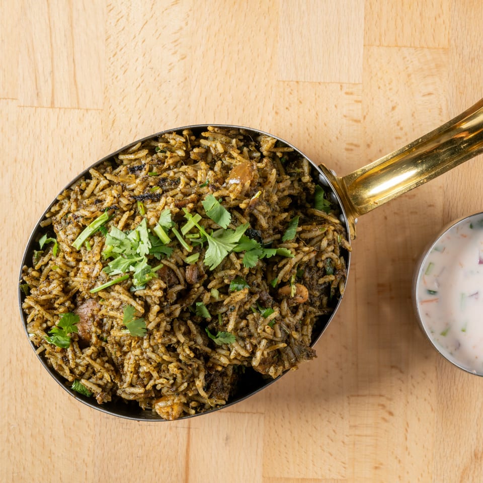 Overhead view of green rice pilaf with cilantro in a brass pan, next to a small bowl of raita.